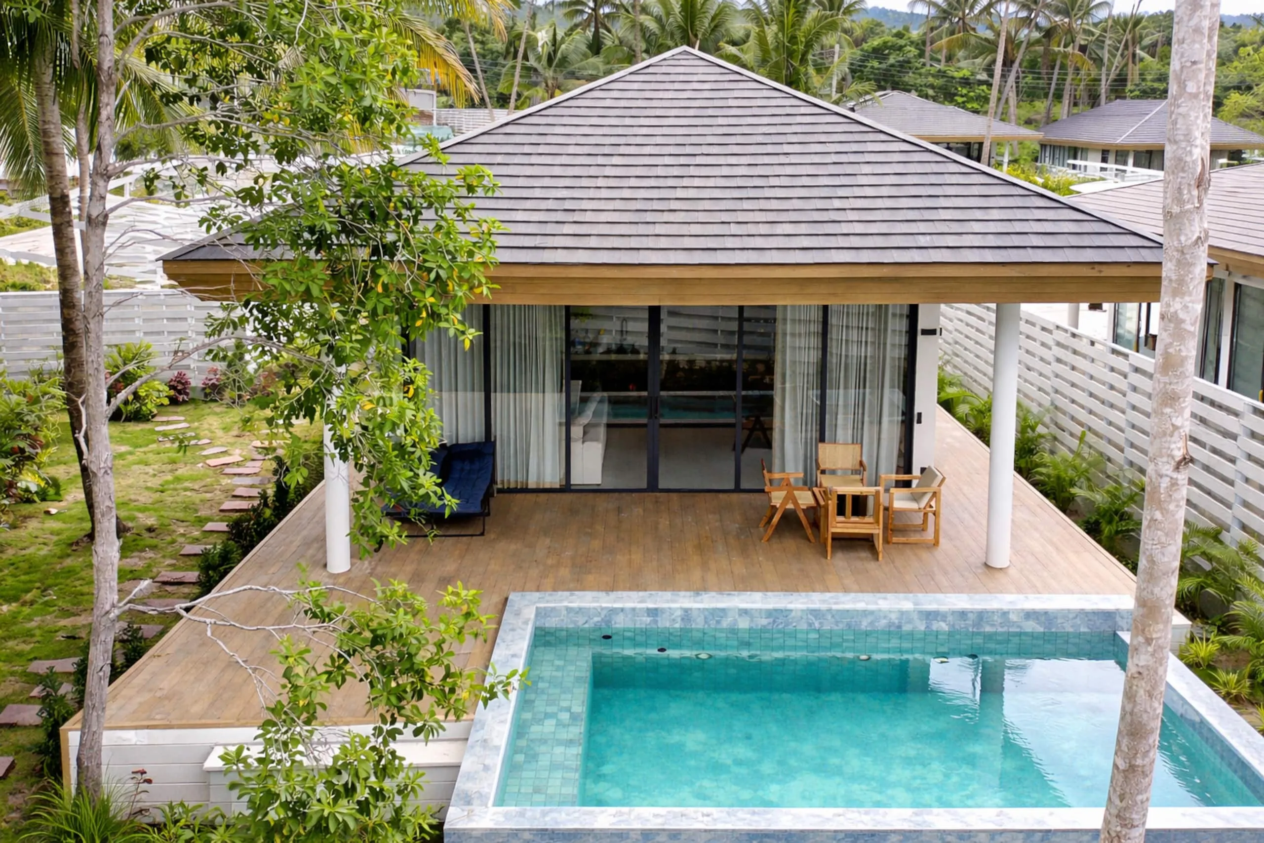 White Lotus Villa II — daytime view over the villa's hipped shingle roof, terrace and turquoise private pool, with palms and green hills behind.